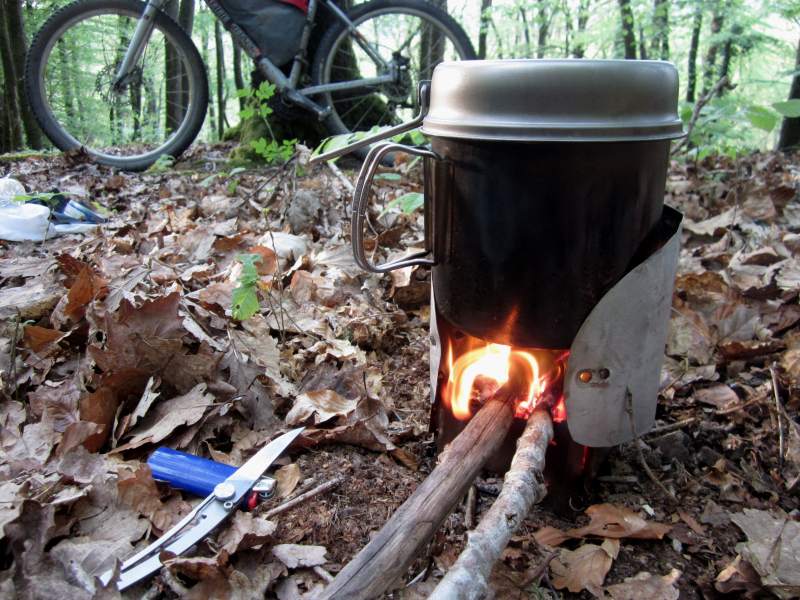 Cuisine de bivouac avec une popotte qui chauffe sur un petit réchaud à bois. 2 petites branches alimentent le mini feu de camps. Les feuilles mortes proches ne risque pas de brûler. La gamelle et le réchaud très léger sont transportées dans une des sacoches du vélo VTT Gravel en arriére-plan.